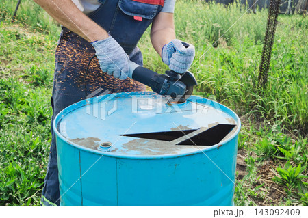 Person using an angle grinder to cut top of blue metal barrel outdoors Person using an angle grinder to cut top of blue metal barrel outdoors 143092409