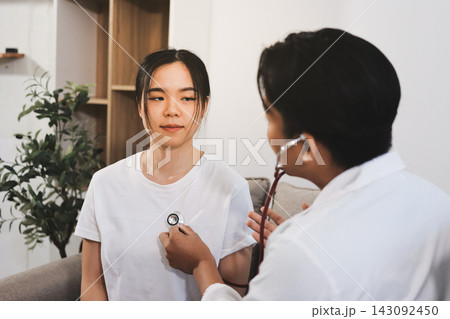 A young male doctor uses a stethoscope to examine a young female patient seated on a couch indoors. A young male doctor uses a stethoscope to examine a young female patient seated on a couch indoors. 143092450