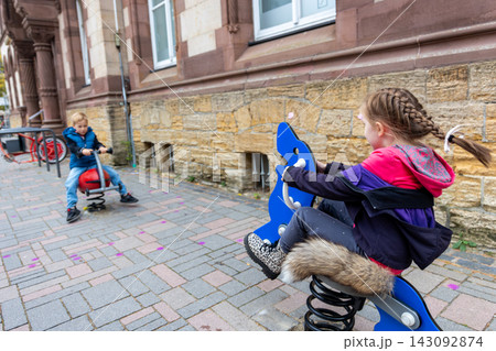 Two children enjoy spring riders on urban sidewalk beside historic stone building, sharing playful outdoor moment. Candid city scene captures childhood, friendship, recreation and active leisure Two children enjoy spring riders on urban sidewalk beside historic stone building, sharing playful outdoor moment. Candid city scene captures childhood, friendship, recreation and active leisure 143092874