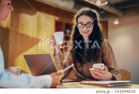 Delighted curly haired woman in glasses sips coffee and checks her smartphone at a wooden table with a colleague Delighted curly haired woman in glasses sips coffee and checks her smartphone at a wooden table with a colleague 143094630