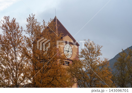 Vintage clock tower surrounded by autumn trees. Old architectural tower with a large dial clock among golden fall foliage against a cloudy sky and mountains. Vintage clock tower surrounded by autumn trees. Old architectural tower with a large dial clock among golden fall foliage against a cloudy sky and mountains. 143096701