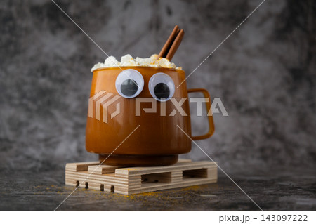 Seasonal drink cacao coffee with whipped cream and cinnamon sticks. Cozy orange cup with funny face eyes halloween concept Thanksgiving and Hello Fall Halloween concept. Celebrating autumn holidays Seasonal drink cacao coffee with whipped cream and cinnamon sticks. Cozy orange cup with funny face eyes halloween concept Thanksgiving and Hello Fall Halloween concept. Celebrating autumn holidays 143097222
