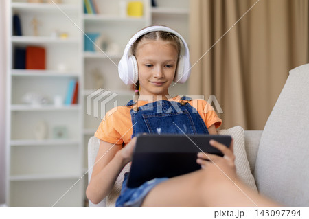 Teen girl wearing headphones relaxing on sofa at home while using tablet Teen girl wearing headphones relaxing on sofa at home while using tablet 143097794