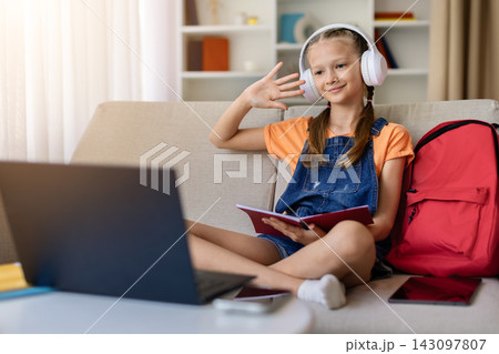 Teen girl in headphones waving toward laptop during online class at home Teen girl in headphones waving toward laptop during online class at home 143097807
