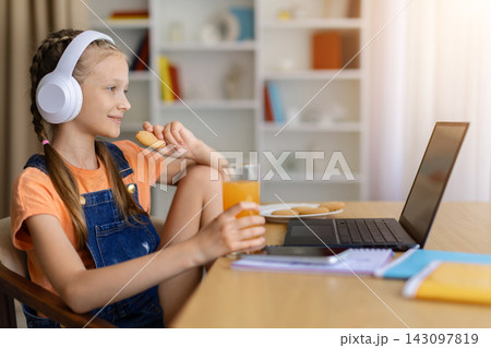 Cheerful preteen girl in headphones eating snacks while watching laptop at home Cheerful preteen girl in headphones eating snacks while watching laptop at home 143097819
