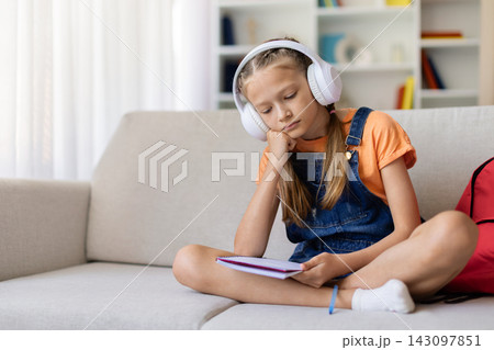 Bored teen girl in headphones sitting on sofa with notebook at home Bored teen girl in headphones sitting on sofa with notebook at home 143097851