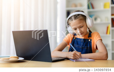 Teen girl wearing headphones writing notes in notebook beside laptop at home Teen girl wearing headphones writing notes in notebook beside laptop at home 143097854