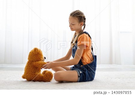 Preteen girl sitting on carpet at home holding paw of teddy bear Preteen girl sitting on carpet at home holding paw of teddy bear 143097855