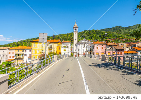 A picturesque view of Kanal Bridge spans the Soca River in Slovenia. Colorful buildings line the street, offering a charming atmosphere against a clear blue sky. 143099548