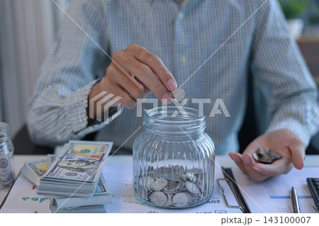 Businessman at working desk putting coins into a glass jar for saving money. Financial planning and saving money concept. Businessman at working desk putting coins into a glass jar for saving money. Financial planning and saving money concept. 143100007