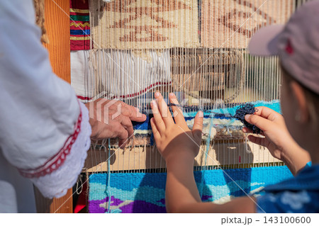 Child Learning Traditional Carpet Weaving at Loom in Kotel, Bulgaria. Child touches yarn while learning traditional carpet weaving from adult at wooden loom during outdoor cultural event 143100600