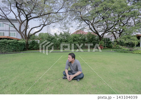 Asian man sits cross legged on a green lawn beneath large trees. He looks away while relaxing in a quiet garden. 143104039