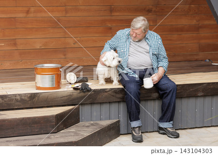 Man relaxes with his dog beside a partially restored wooden terrace, oil treatment can and sanding tools, illustrating outdoor deck maintenance and renovation. 143104303