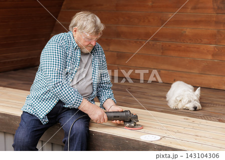 Senior homeowner uses an electric sander to restore outdoor wooden deck boards while a dog rests nearby, illustrating DIY maintenance and refinishing. 143104306