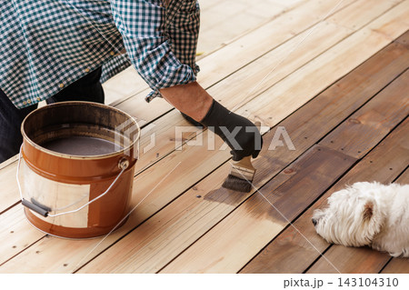 A gloved worker applies protective oil with a brush to wooden terrace decking, illustrating outdoor maintenance, surface treatment, and renovation for residential property care. 143104310