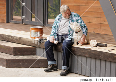 Senior homeowner sits with his dog on a wooden deck beside sanding equipment and a paintbrush during an outdoor restoration project. Senior homeowner sits with his dog on a wooden deck beside sanding equipment and a paintbrush during an outdoor restoration project. 143104311