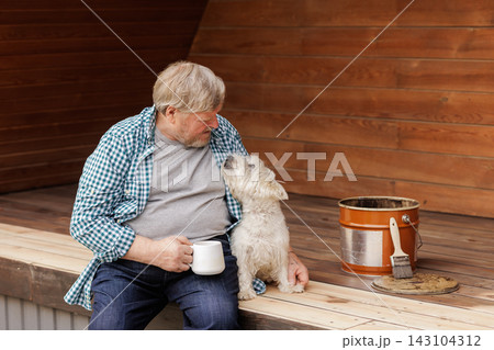 Senior homeowner relaxes with his dog on a wooden terrace beside a brush and bucket, illustrating deck maintenance, outdoor living, and wood treatment. Senior homeowner relaxes with his dog on a wooden terrace beside a brush and bucket, illustrating deck maintenance, outdoor living, and wood treatment. 143104312