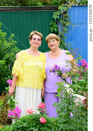Authentic portrait of two smiling senior russian sisters 80 and 75 years old with flower bouquets standing in a summer garden in Russia 143106976