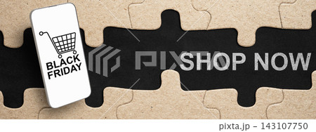 A puzzle concept with cardboard jigsaw pieces revealing a black background with white text saying SHOP NOW and a smartphone graphic with a shopping cart icon. Concept of Black Friday Sale 143107750