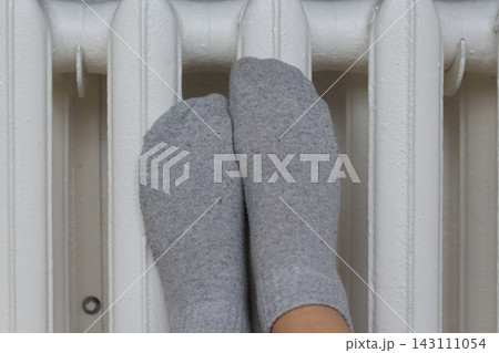 Person in gray knit socks warming up cold feet on a white central heating unit Person in gray knit socks warming up cold feet on a white central heating unit 143111054