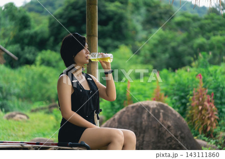 Woman in black sleeveless outfit sitting outdoors drinking water from clear plastic bottle in green nature setting Woman in black sleeveless outfit sitting outdoors drinking water from clear plastic bottle in green nature setting 143111860