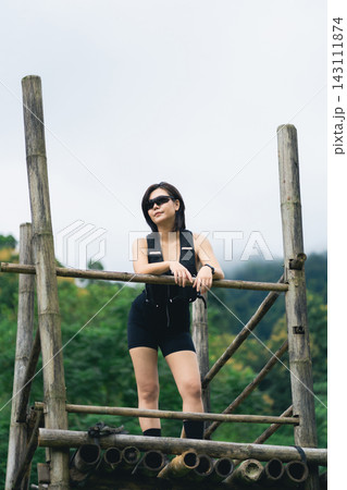 Young confident woman in black outfit and sunglasses standing on rustic wooden platform outdoors with green forest background Young confident woman in black outfit and sunglasses standing on rustic wooden platform outdoors with green forest background 143111874