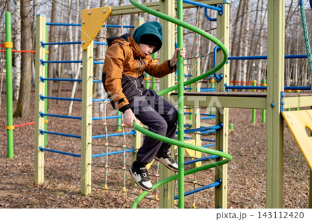 preteen boy in an orange jacket climbing up a green spiral ladder on a children's playground structure outdoors in autumn or spring. preteen boy in an orange jacket climbing up a green spiral ladder on a children's playground structure outdoors in autumn or spring. 143112420