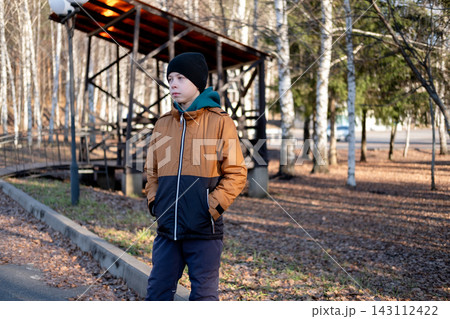 young caucasian boy with hands in pockets wearing a brown winter jacket standing on a park path near a wooden alcove or gazebo. 143112422