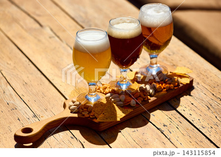 Three craft beer glasses with snacks on wooden board in sunny outdoor bar. 143115854