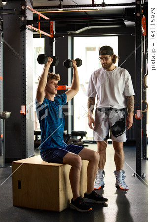 Young athlete doing seated dumbbell shoulder press with trainer in gym. Young athlete doing seated dumbbell shoulder press with trainer in gym. 143115979