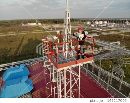 Male worker in red helmet inspects tower lights on industrial mast, focused in hazardous setting. This aerial industrial view shows maintenance worker on raised platform beside power infrastructure. Male worker in red helmet inspects tower lights on industrial mast, focused in hazardous setting. This aerial industrial view shows maintenance worker on raised platform beside power infrastructure. 143117093
