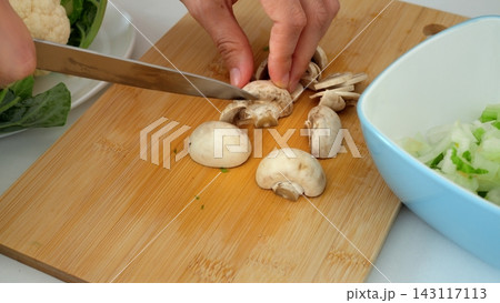 Chef practices knife safety while slicing button mushroom on bamboo board in bright kitchen, demonstrating safe finger positioning alongside fresh cauliflower, celery, and salad greens. 143117113