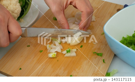 Cook slices fresh white onion using kitchen knife on bamboo cutting board during culinary food prep. Focused hands chop fresh vegetables carefully beside bowl of salad greens and cauliflower. Cook slices fresh white onion using kitchen knife on bamboo cutting board during culinary food prep. Focused hands chop fresh vegetables carefully beside bowl of salad greens and cauliflower. 143117115