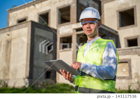 Construction professional using tablet at unfinished building site while wearing hard hat and safety vest. Architectural services construction technology and mobile project management Construction professional using tablet at unfinished building site while wearing hard hat and safety vest. Architectural services construction technology and mobile project management 143117536