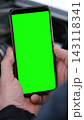 Male hands holding digital smart phone mockup template with green screen chroma key over an open car hood with engine. Close up of driver user cell phone display near automobile motor. Vertical photo. 143118341
