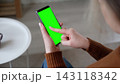 Over the shoulder view of woman holding smartphone vertically and touching green screen mockup chroma key display. Girl tapping on mobile phone template mockup blank screen inside a cozy room 143118342