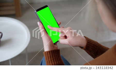 Over the shoulder view of woman holding smartphone vertically and touching green screen mockup chroma key display. Girl tapping on mobile phone template mockup blank screen inside a cozy room Over the shoulder view of woman holding smartphone vertically and touching green screen mockup chroma key display. Girl tapping on mobile phone template mockup blank screen inside a cozy room 143118342