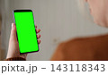 Over the shoulder view of young woman holding smartphone vertically with green screen mockup chroma key blank display. Female user looking at phone template mockup screen inside cozy room at home 143118343