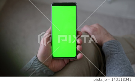 Top view of man holding smartphone vertically with green screen mockup chroma key blank display. Male user sitting and looking at mobile phone template mockup surface inside cozy room at home Top view of man holding smartphone vertically with green screen mockup chroma key blank display. Male user sitting and looking at mobile phone template mockup surface inside cozy room at home 143118345