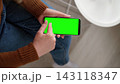 Top view of woman holding smartphone horizontally and touching green screen mockup chroma key display. Girl sitting on chair and using mobile phone template mockup blank surface next to table 143118347
