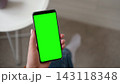 Top view of woman holding smartphone with vertical green screen chroma key inside cozy room. Girl sitting on sofa using mobile phone template mockup display next to a coffee cup on white round table 143118348