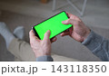 Close up of male hands holding smartphone horizontally with green screen mockup display inside cozy room. Man playing mobile game or watching video using chroma key blank template smart phone 143118350