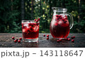 An ice-cold glass of cranberry juice with condensation next to a glass pitcher and scattered fresh cranberries on dark wood. 143118647