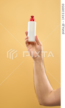 Hand holding white skincare bottle with red pump on soft yellow background. 143118950