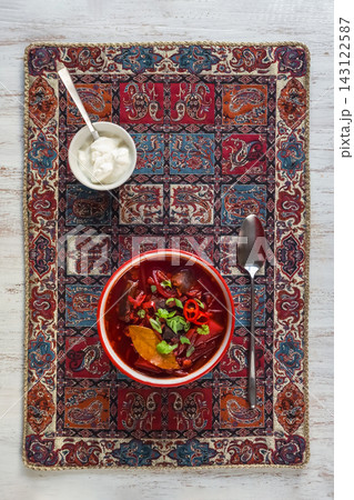 Traditional beetroot soup borscht with sour cream on an ornamental mat. Top view flat lay of red beet soup in a bowl served with metal spoon and sour cream on an ethnic pattern background. Traditional beetroot soup borscht with sour cream on an ornamental mat. Top view flat lay of red beet soup in a bowl served with metal spoon and sour cream on an ethnic pattern background. 143122587