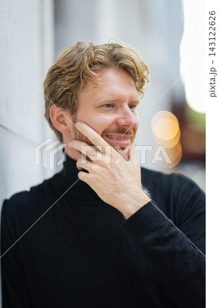 Man in black turtleneck smiles while touching his face in an urban setting with blurred background lights Man in black turtleneck smiles while touching his face in an urban setting with blurred background lights 143122626