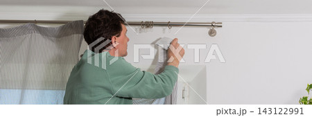 A man is hanging a curtain on the rod after washing it. It is spring, and he is doing household chores in a room with a window and some greenery, banner 143122991