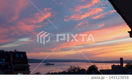 Pink and orange sunset clouds sweep over the sea while dark buildings and a roof edge frame the coastal view. The image symbolizes peaceful endings, transition, and harmony between city and nature. 143123058