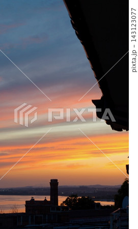 Vertical coastal sunset shows orange light near the horizon beneath layered pink and blue clouds framed by a dark roof edge. The scene conveys closure, stillness, and the contrast between architecture Vertical coastal sunset shows orange light near the horizon beneath layered pink and blue clouds framed by a dark roof edge. The scene conveys closure, stillness, and the contrast between architecture 143123077