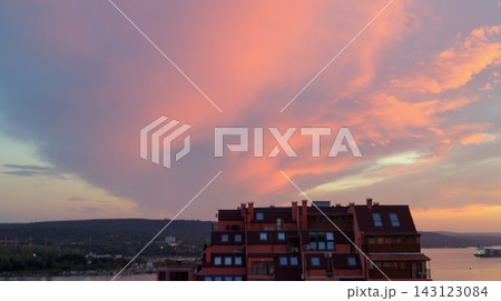 A large coral pink cloud glows above seaside apartment buildings beneath a soft blue and orange sunset sky. The image expresses contrast between urban life and the calm beauty of nature. A large coral pink cloud glows above seaside apartment buildings beneath a soft blue and orange sunset sky. The image expresses contrast between urban life and the calm beauty of nature. 143123084